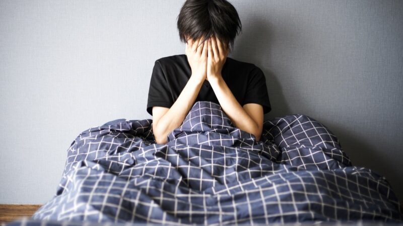 朝、ベッドの上で顔を覆って座り込む男性